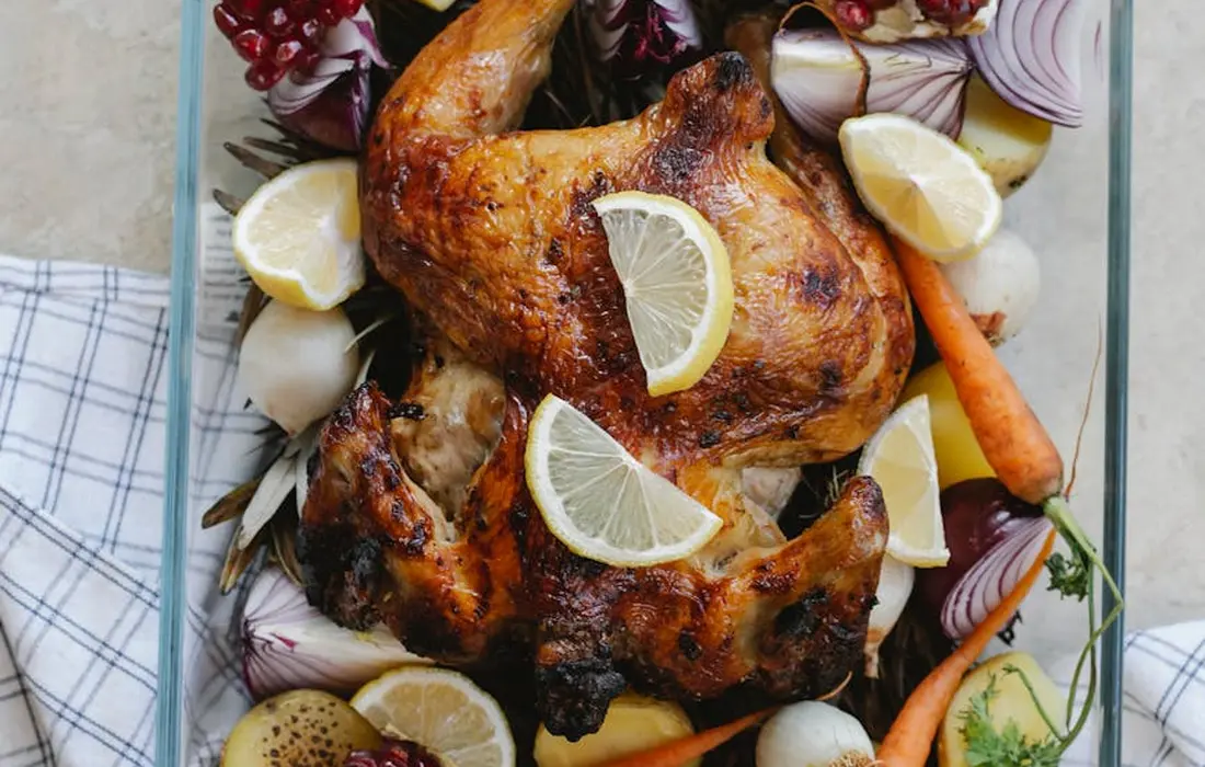 A roasted chicken on a baking sheet garnished with lemon wedges and surrounded by onions, carrots, and herbs.