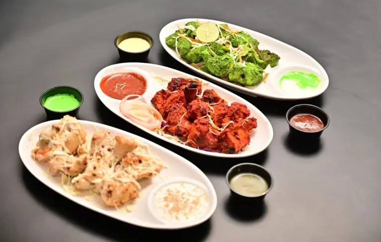 Three white plates on a dark surface containing fried chicken pieces, a green salad, and assorted dipping sauces served in small cups.