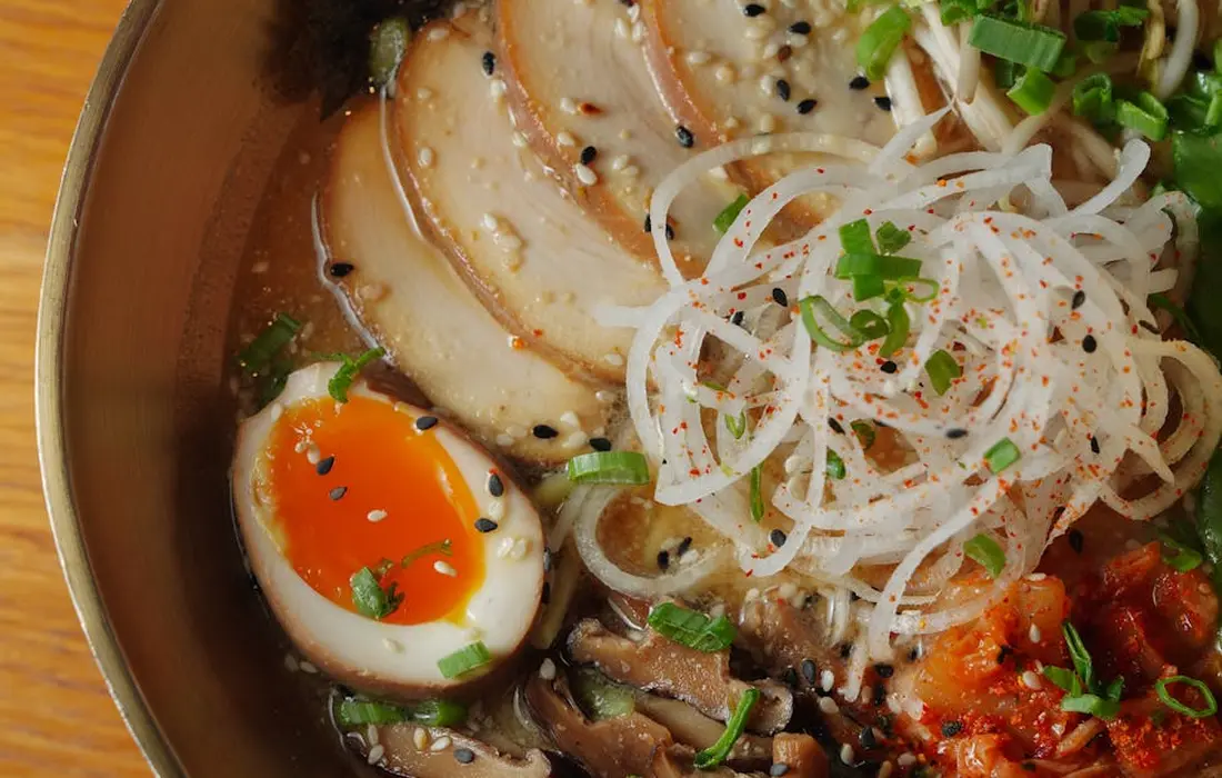 Close-up of a bowl with sliced meat, a halved soft-boiled egg, and thin ribbons of onion, garnished with green onions and sesame seeds in a rich broth.