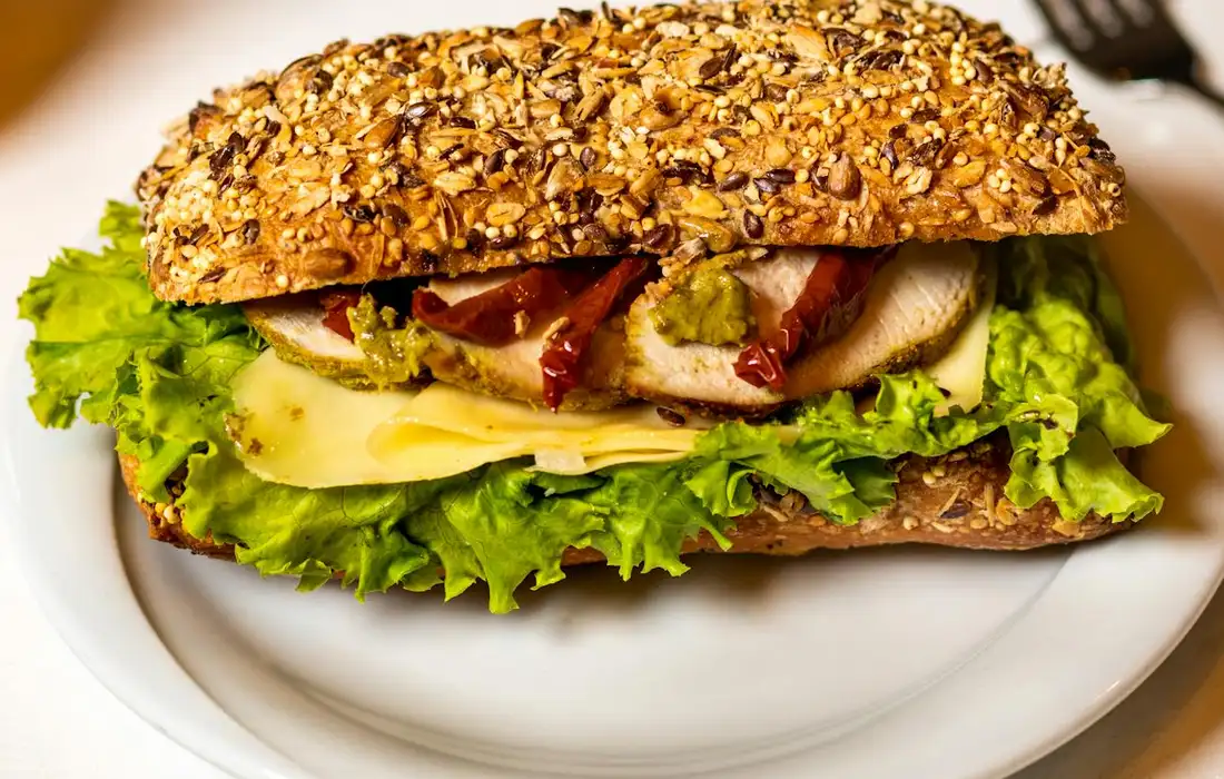 Close-up of a seeded whole-grain sandwich on a white plate, filled with lettuce, cheese, and deli meat.