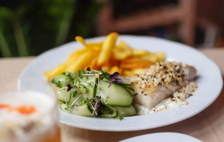 Plate of sesame-crusted fish fillet with cucumber salad microgreens and French fries on a white plate.