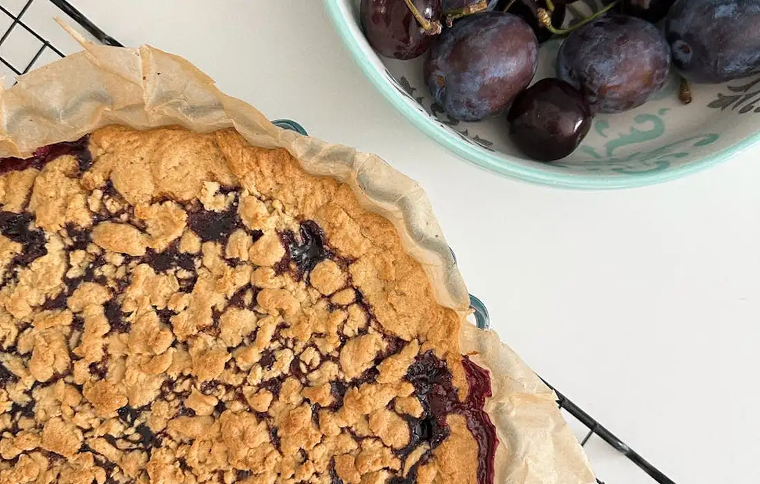 Plum pie with a lattice crust placed on a white surface, beside a bowl of fresh plums