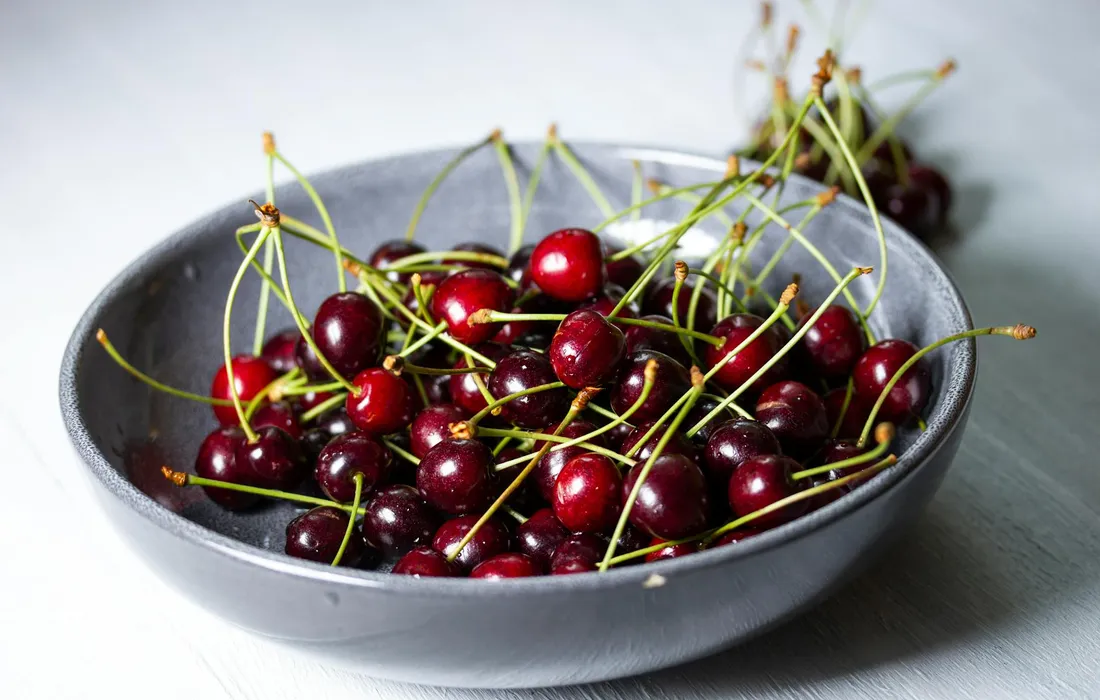 A bowl of fresh red cherries with stems on a light surface