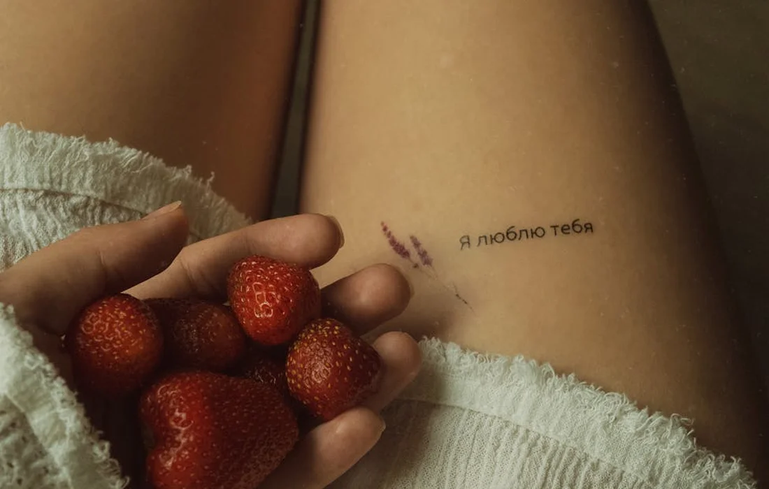Close-up of a hand holding fresh strawberries beside a leg with a small tattoo that reads 'Я люблю тебя'.