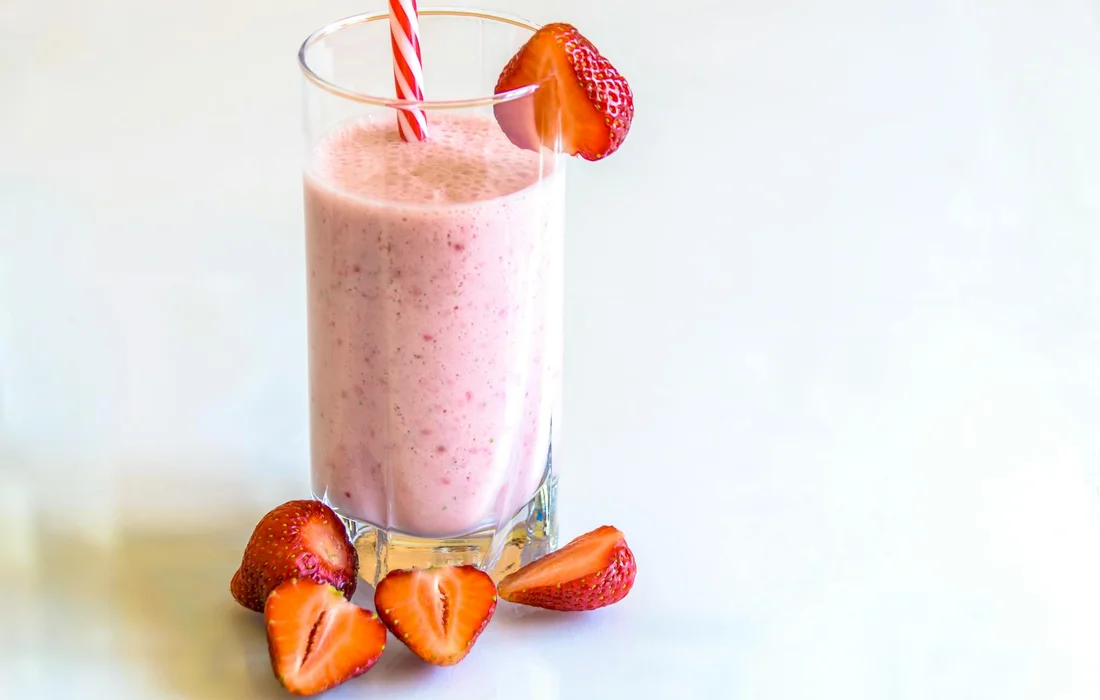 A tall glass of pink strawberry smoothie with a red-and-white striped straw, garnished with a strawberry slice; fresh strawberries sit nearby on a light surface.