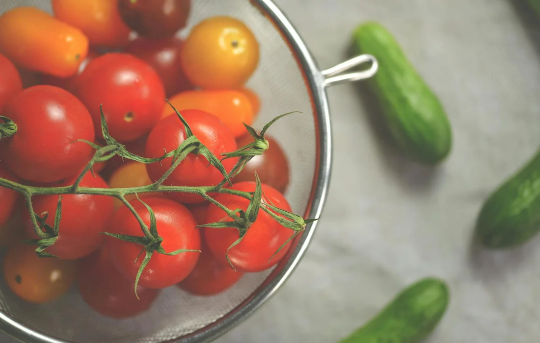 Cherry tomatoes in a metal colander with cucumbers resting on a light countertop.