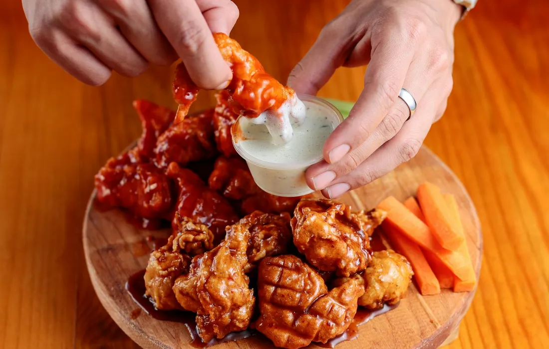 Hands reaching for a piece of fried chicken from a wooden plate, with a bowl of white dipping sauce and carrot sticks on the side.