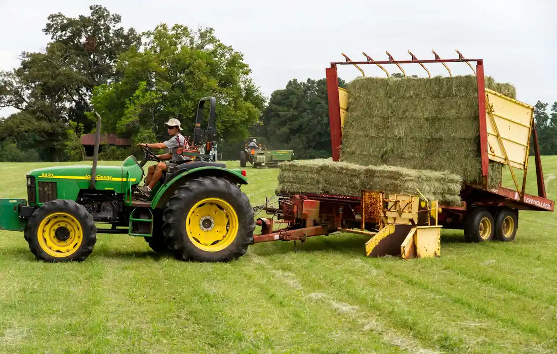 Green tractor pulling a trailer loaded with rectangular hay bales across a grassy field.