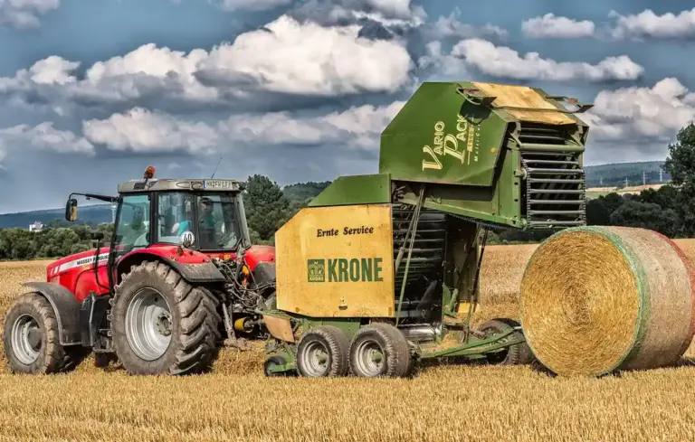 Red tractor with a green hay baler in a harvested field, with large round hay bales in the background