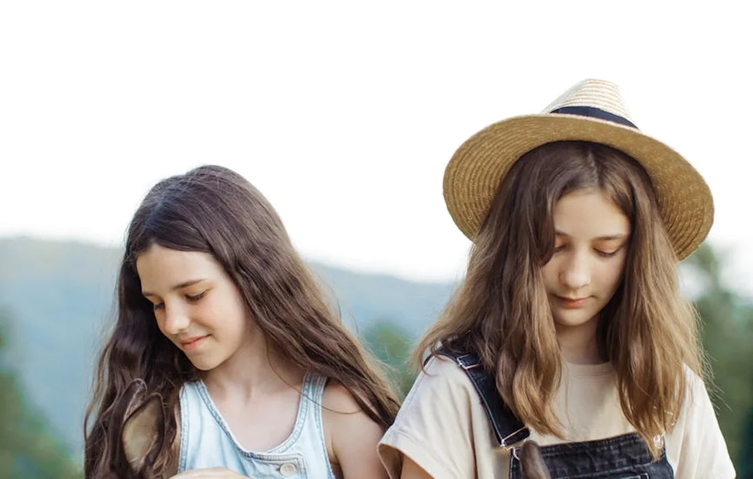 Two young girls standing outdoors, looking down at something, with a blurred green landscape in the background