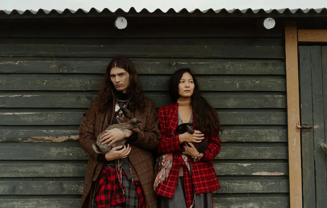 Two people standing in front of a weathered wooden wall, each holding a rabbit.