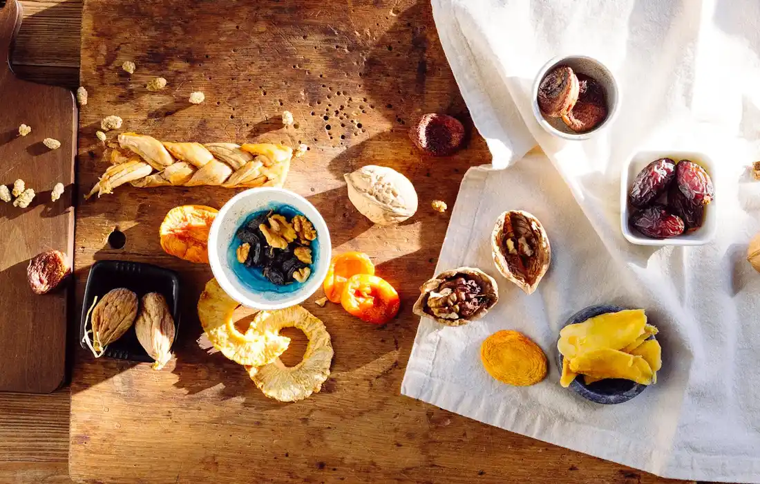 Assorted dried fruits and nuts scattered on a wooden surface, including raisins, apricots, mango slices, and nuts, with a white napkin nearby.
