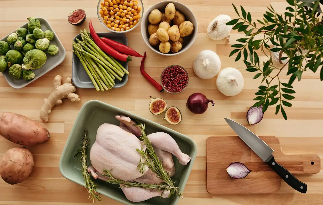 Raw chicken on a green tray amid onions, garlic, peppers, and herbs on a wooden cutting board with a knife nearby.