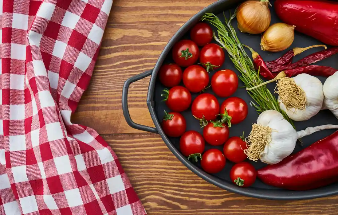 Tray of fresh vegetables including cherry tomatoes, red peppers, onions, garlic, and rosemary on a wooden table with a red-and-white checkered cloth.