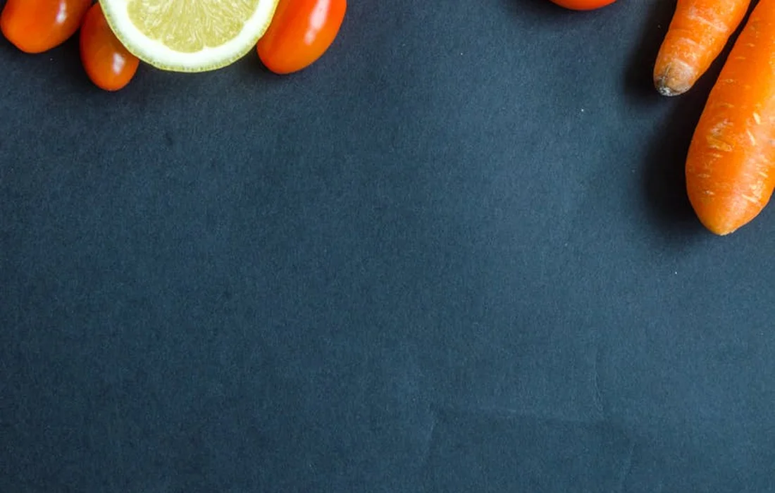 Assorted vegetables on a dark slate background, featuring carrots and a lemon slice.