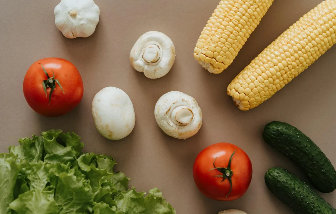Assorted fresh vegetables on a brown background, including romaine lettuce, tomatoes, corn on the cob, white mushrooms, cucumbers, and garlic.