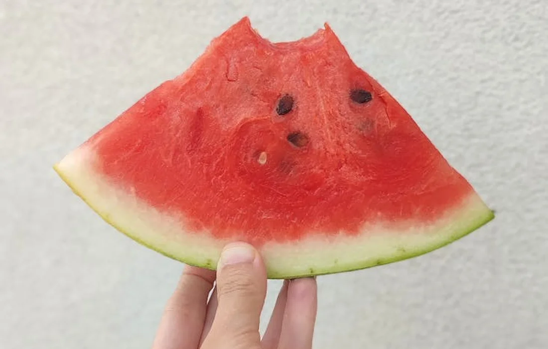 A hand holding a triangular slice of watermelon rind against a light, neutral background.