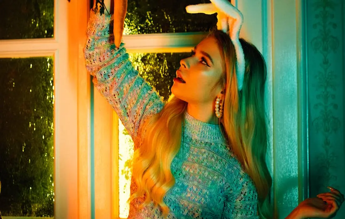 Portrait of a young woman with long blonde hair wearing a light blue knit sweater, standing by a window with warm, colorful lighting.
