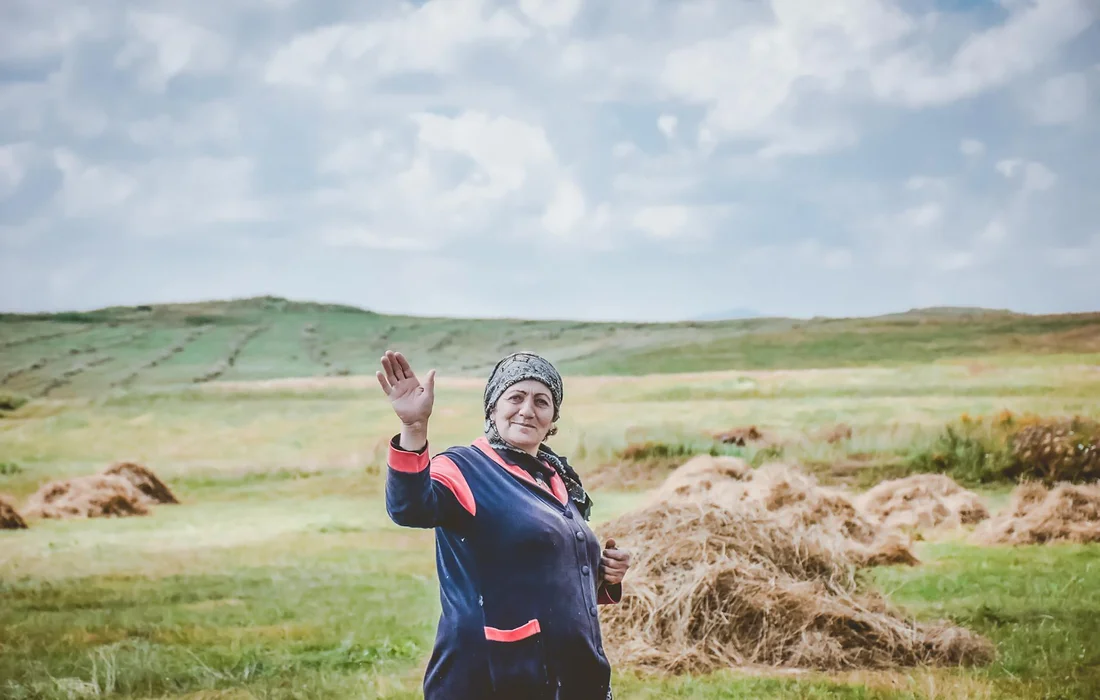 An older woman in a blue jacket and headscarf stands in a grassy field with hay piles, waving at the camera.