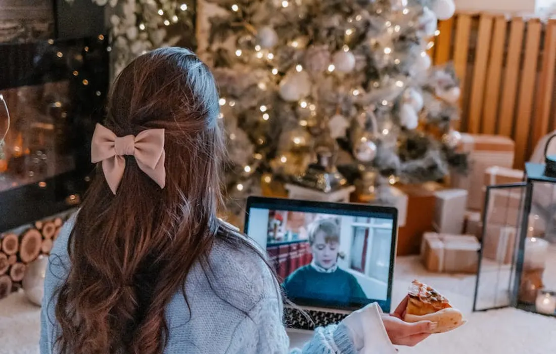 A person with a pink bow in her hair sits in a festively decorated living room, watching a laptop screen while holding a pastry.